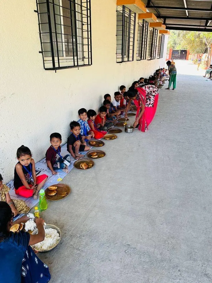 School children seated in a row being served mid-day meals by volunteers